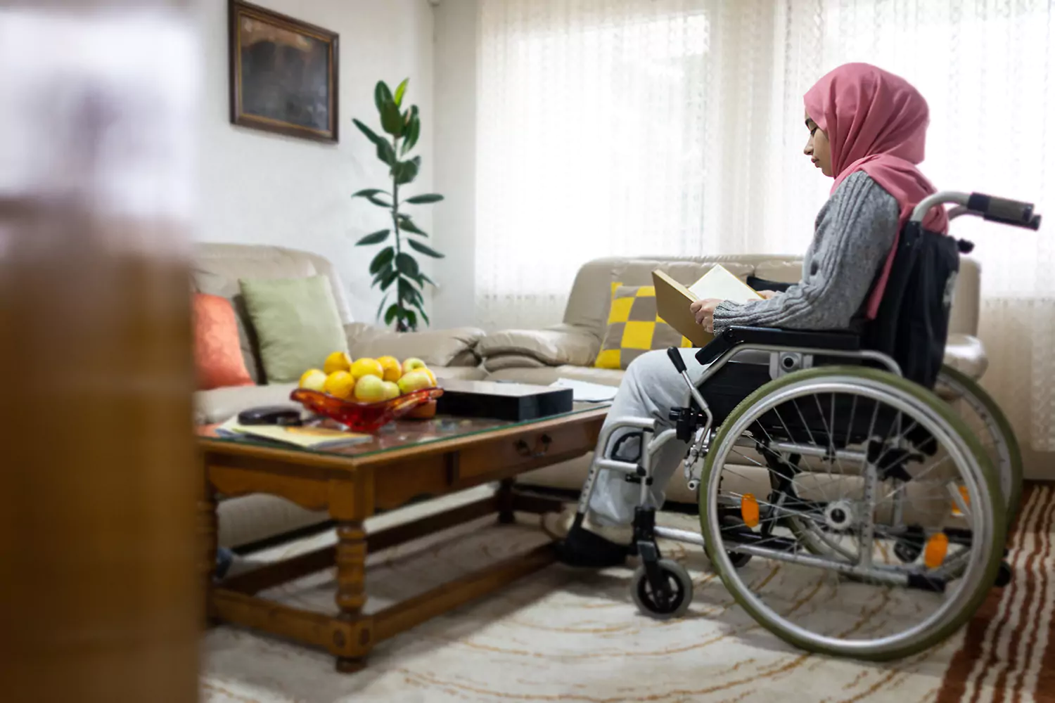 A woman in a wheelchair is reading a book in her living room